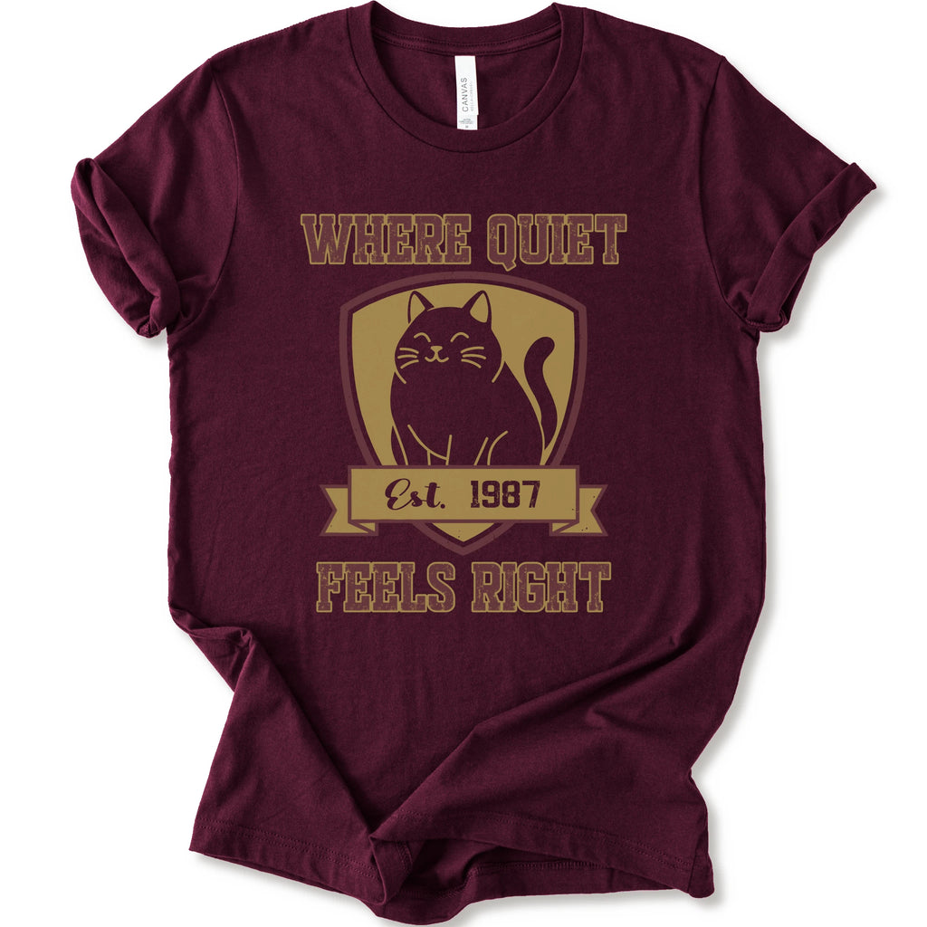 A maroon short-sleeve t-shirt with a vintage-style graphic on the front featuring a black cat inside a gold shield and the text 'WHERE QUIET FEELS RIGHT Est. 1987'.