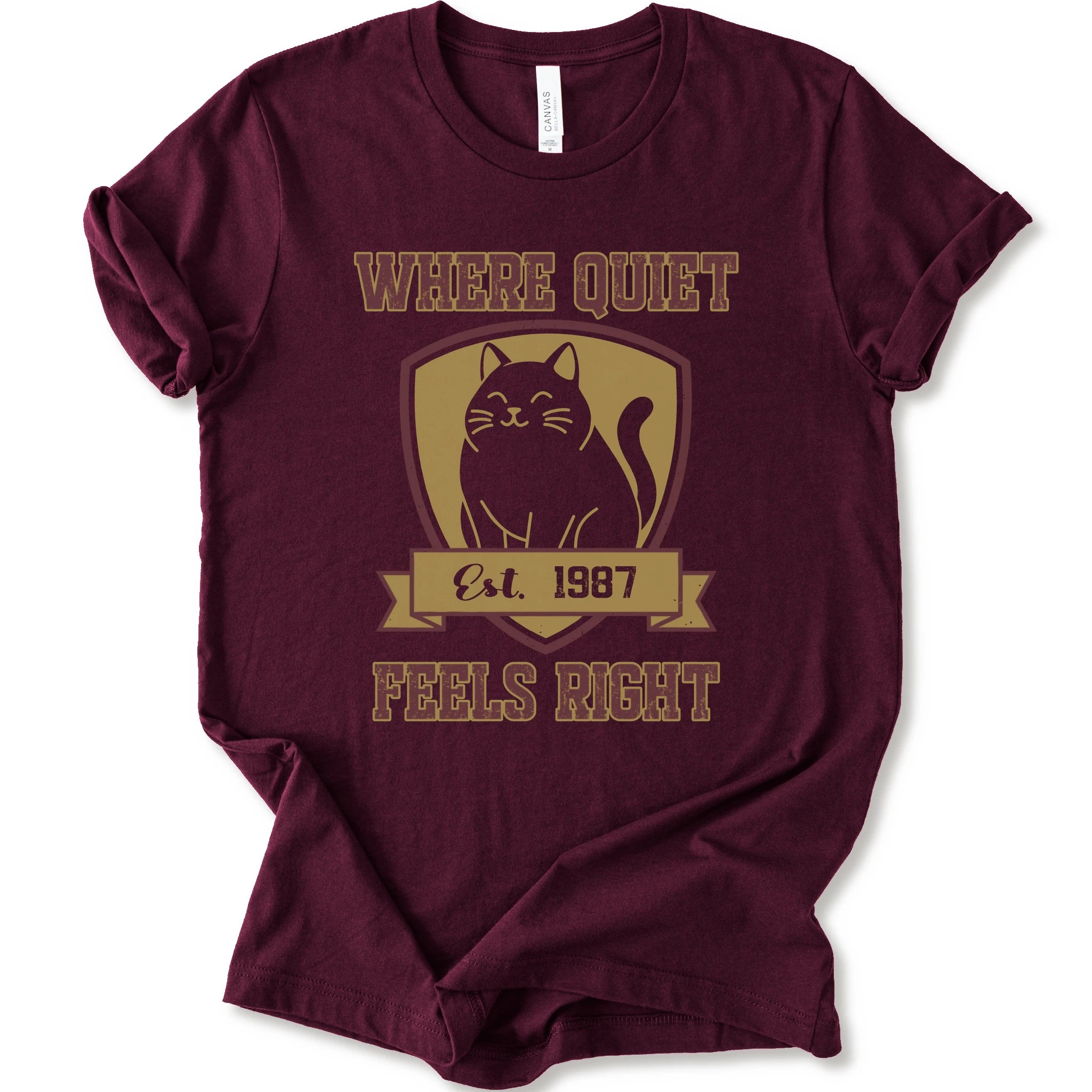 A maroon short-sleeve t-shirt with a vintage-style graphic on the front featuring a black cat inside a gold shield and the text 'WHERE QUIET FEELS RIGHT Est. 1987'.