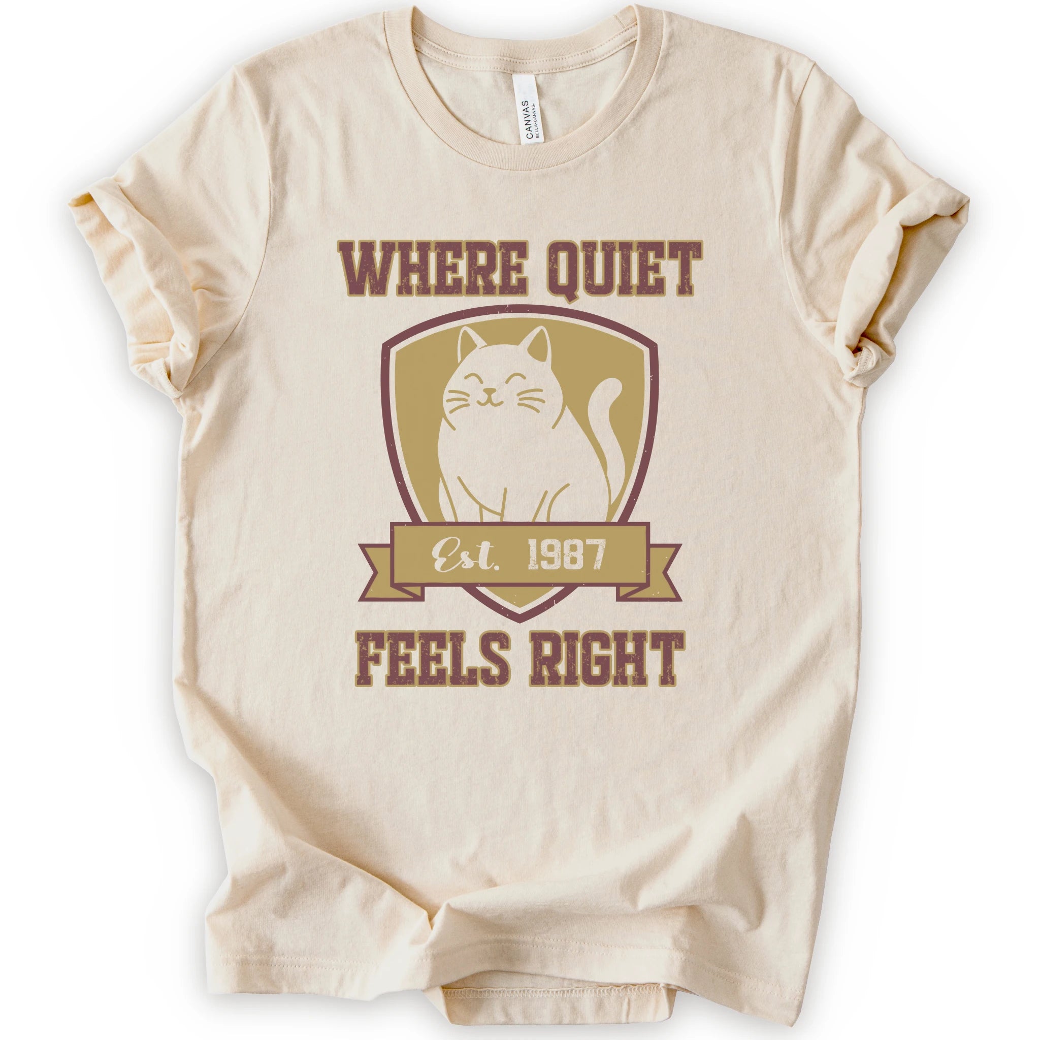 A natural short-sleeve t-shirt with a vintage-style graphic on the front featuring a black cat inside a gold shield and the text 'WHERE QUIET FEELS RIGHT Est. 1987'.