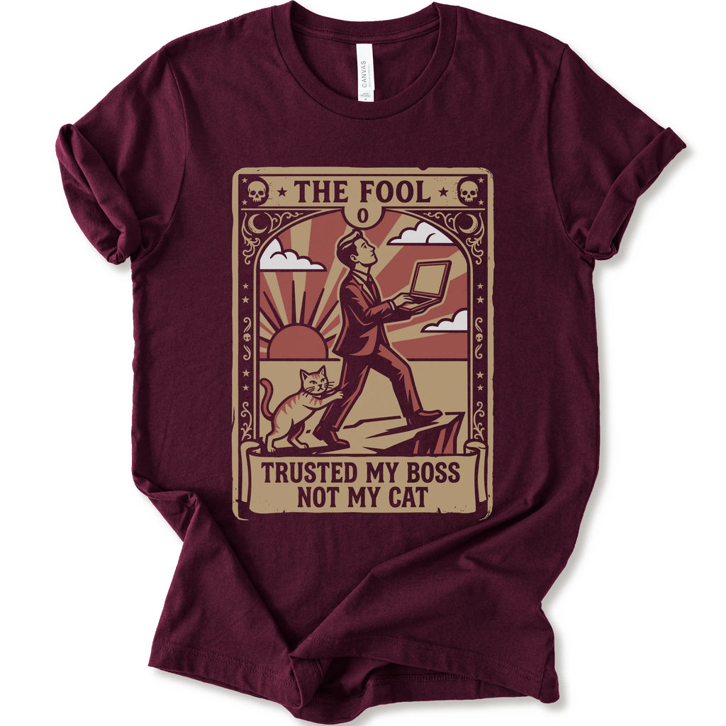 A maroon short-sleeve crewneck t-shirt with a vintage-style graphic print on the front. The graphic is styled like an old tarot card, featuring a man in a suit walking on a cliff while a cat watches, with the text 'THE FOOL' at the top and 'TRUSTED MY BOSS NOT MY CAT' at the bottom.