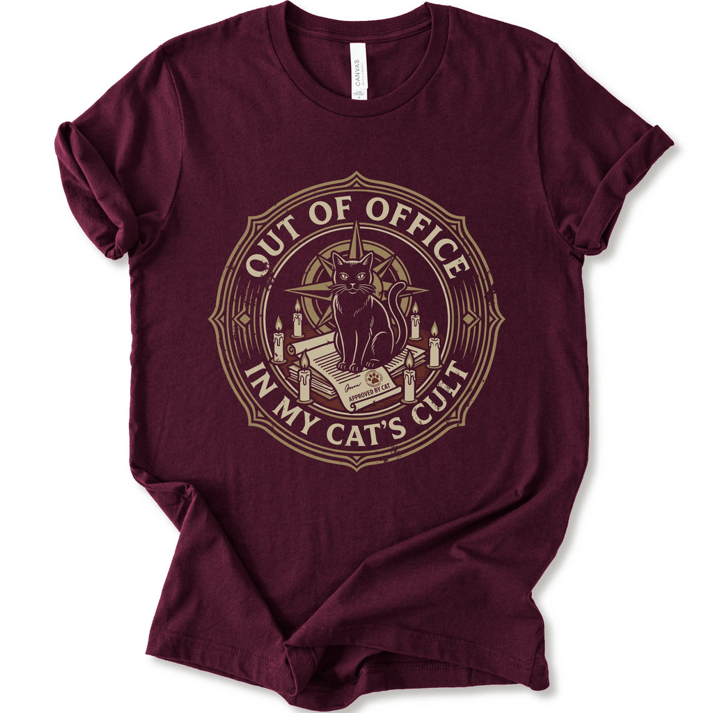 A maroon short-sleeve crewneck t-shirt with a circular graphic on the chest. The graphic features a black cat sitting on an open book surrounded by candles, with the text 'OUT OF OFFICE' and 'IN MY CAT'S CULT' in a vintage serif font.