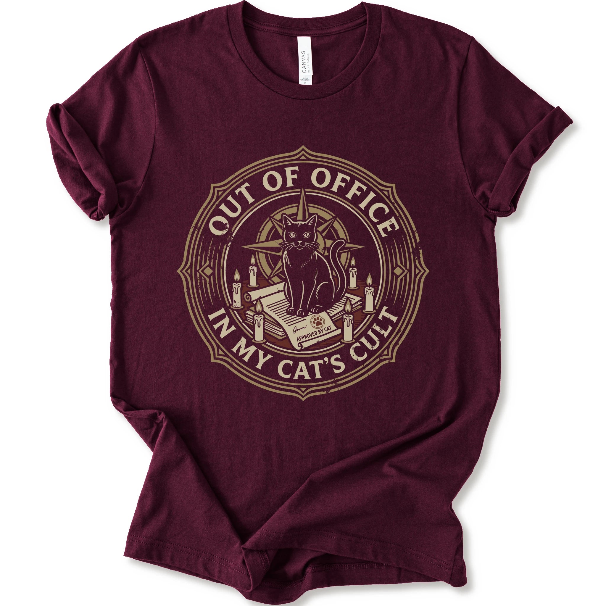 A maroon short-sleeve crewneck t-shirt with a circular graphic on the chest. The graphic features a black cat sitting on an open book surrounded by candles, with the text 'OUT OF OFFICE' and 'IN MY CAT'S CULT' in a vintage serif font.