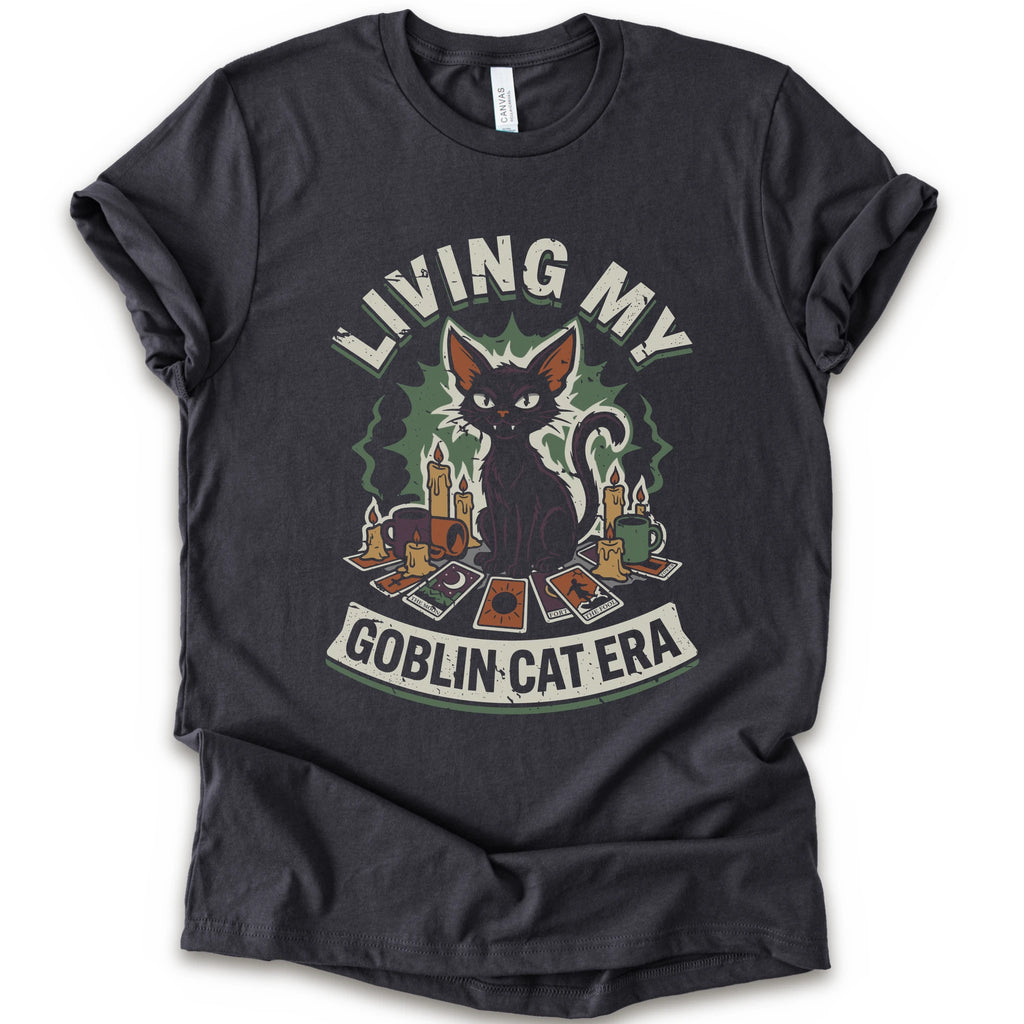 A dark grey short-sleeve t-shirt with a graphic print of a black cat surrounded by candles, cards, and the text 'LIVING MY GOBLIN CAT ERA' in a distressed vintage style.