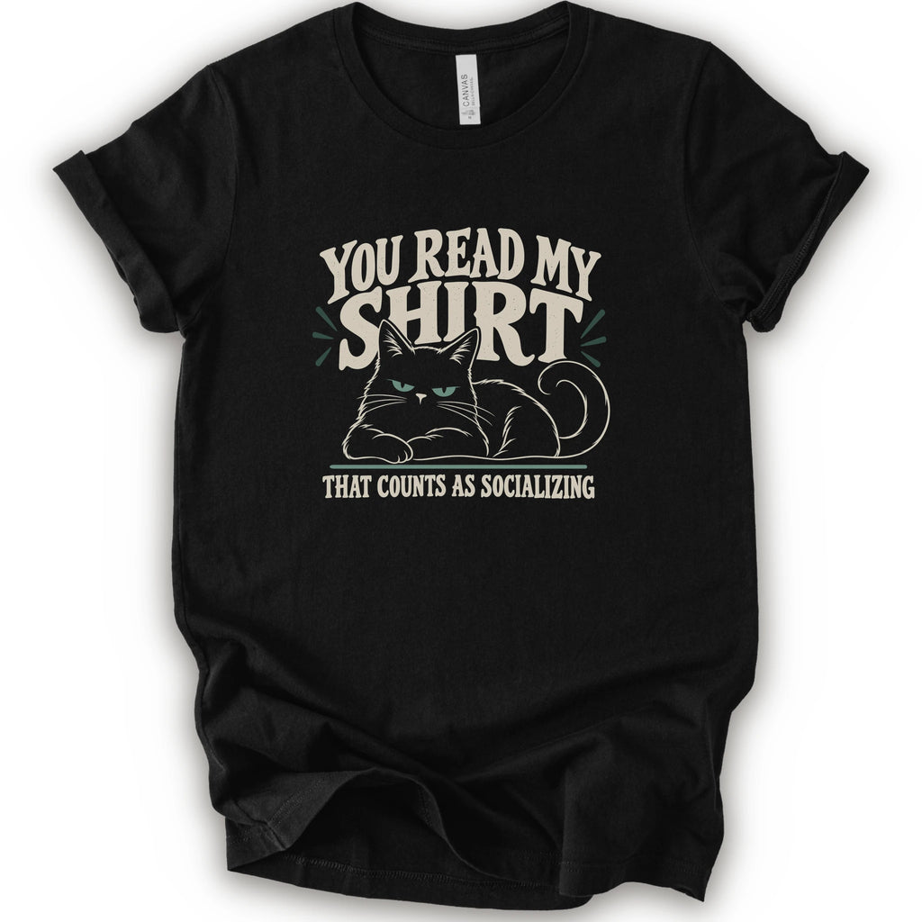 A black short-sleeve crewneck t-shirt with a graphic print of a grumpy cat and the text 'YOU READ MY SHIRT THAT COUNTS AS SOCIALIZING' in cream and light green.