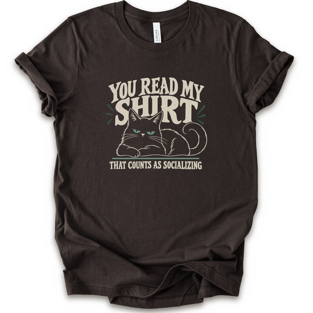 A brown short-sleeve crewneck t-shirt with a graphic print of a grumpy cat and the text 'YOU READ MY SHIRT THAT COUNTS AS SOCIALIZING' in cream and light green.