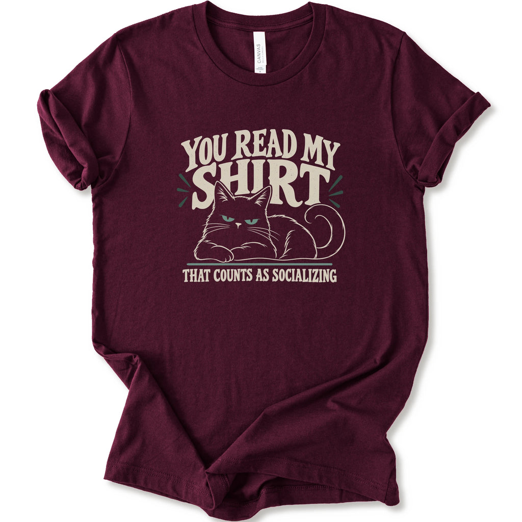 A maroon short-sleeve crewneck t-shirt with a graphic print of a grumpy cat and the text 'YOU READ MY SHIRT THAT COUNTS AS SOCIALIZING' in cream and light green.