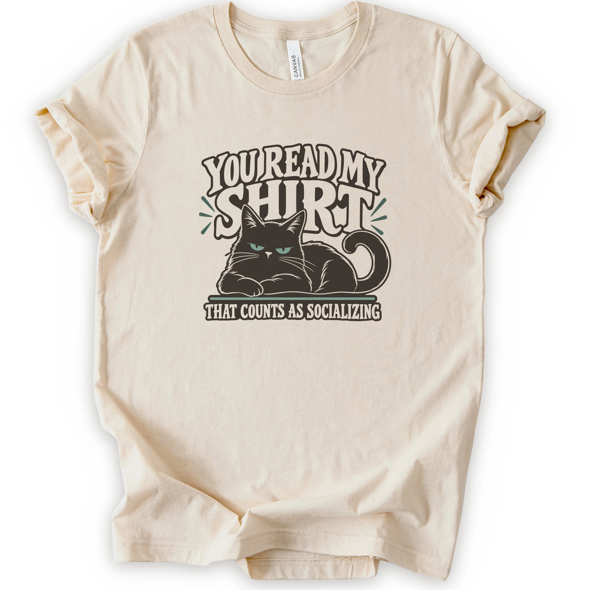 A natural short-sleeve crewneck t-shirt with a graphic print of a grumpy cat and the text 'YOU READ MY SHIRT THAT COUNTS AS SOCIALIZING' in brown and light green.